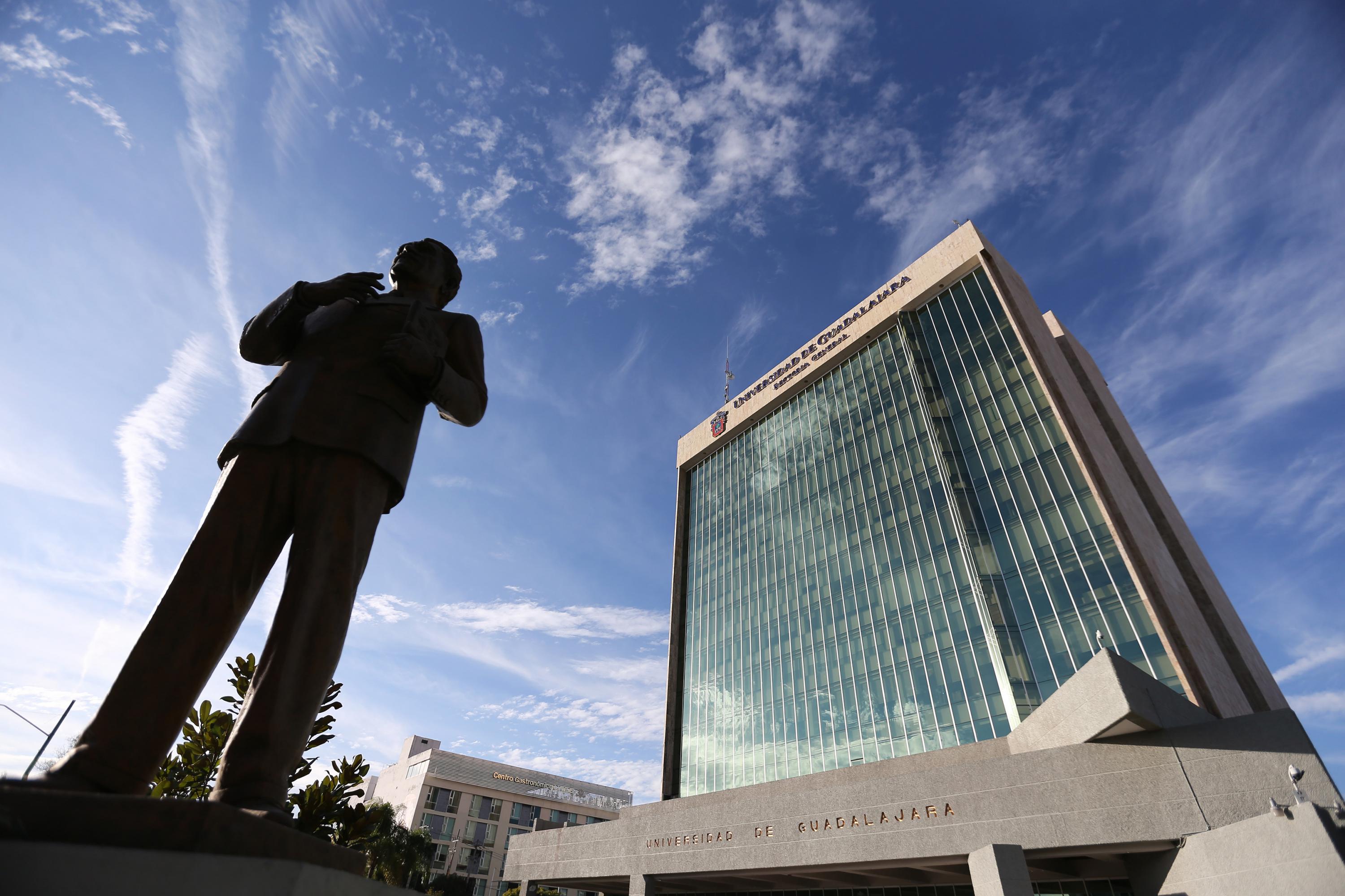 Es una fotografía en la que se muestra el edificio de la Rectoría General de la Universidad de Guadalajara, el cielo está azul y al lado izquierdo aparece la estatua de Constancio Hernández Alvirde